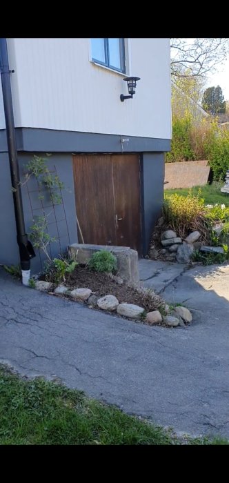 Brunt trähus med vit övervåning som delvis döljer en källartrappa och dörr nedanför, omgivet av växtlighet och stenar.