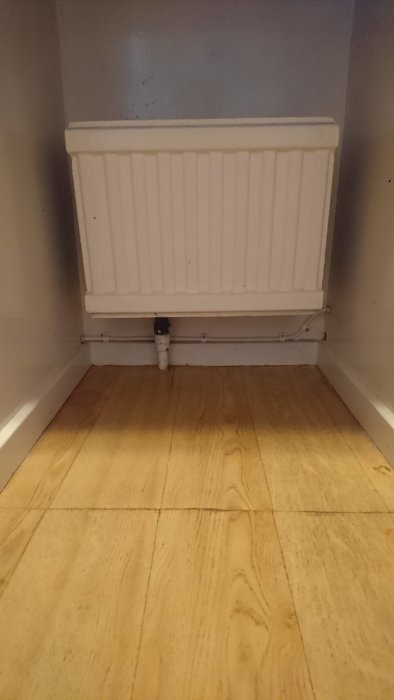 En vit radiator med termostatventil i ett hörn med trägolv.