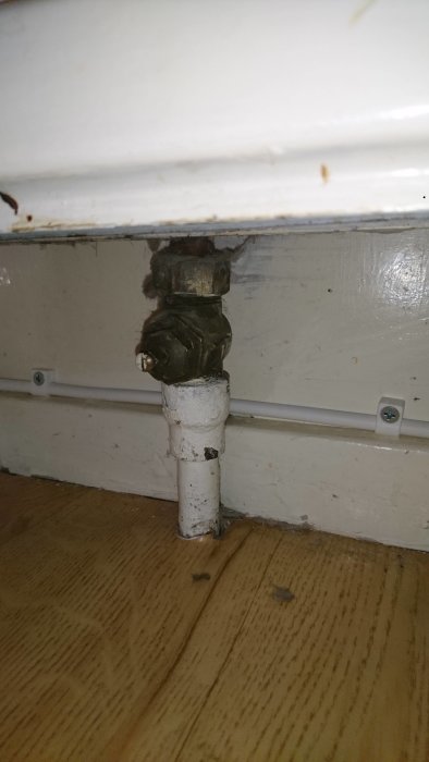 Närbild på en gammal och sliten radiatorventil kopplad till ett vitt rör.