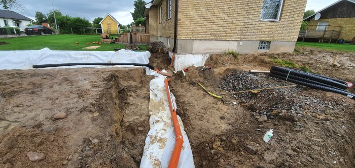 Grävd ränna för kulvert och installationer med synliga elrör, vattenledningar och dränering vid husgrund.