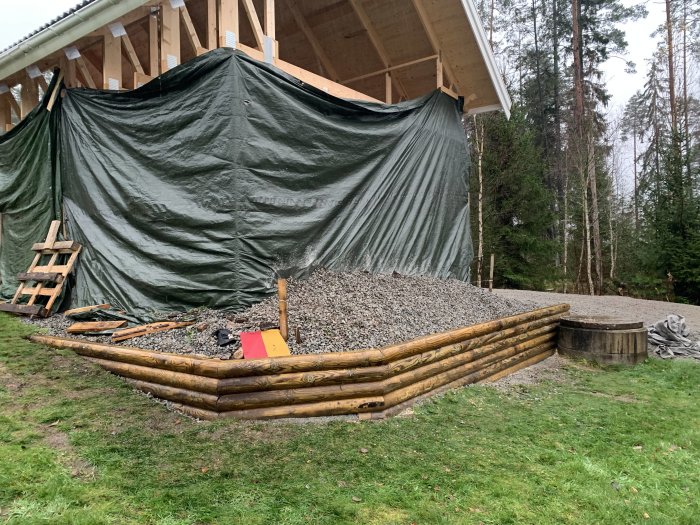 En nyligen byggd stödmur av trästockar med stenbakfyllning vid en byggarbetsplats med täckt byggnad i bakgrunden.