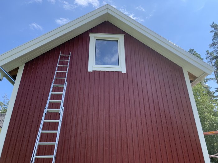 En rödmålad husvägg med vit snickarglädje under taket och en stege lutad mot väggen.