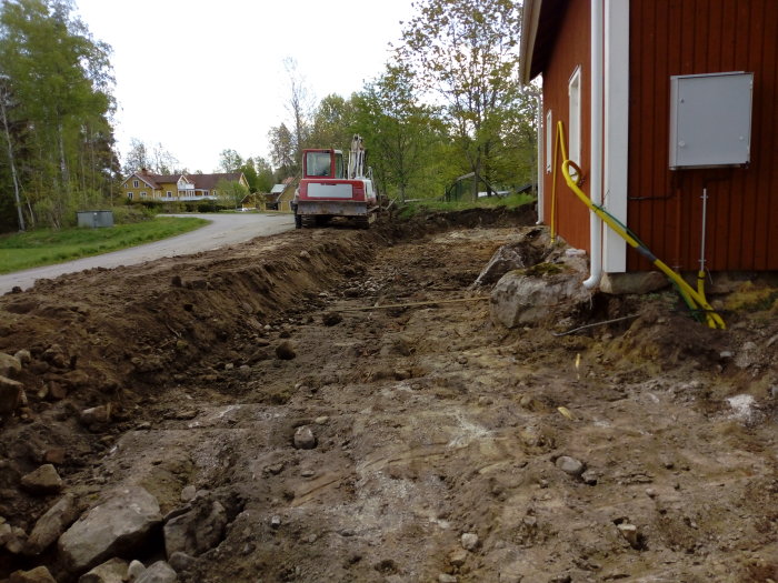 Grävarbete vid grund för byggprojekt, röd grävmaskin och grusig byggarbetsplats nära rött hus.