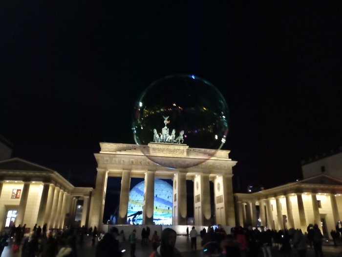 Kvällsbild av Brandenburger Tor i Berlin med en stor reflekterande sfär framför och människor som samlats runtom.