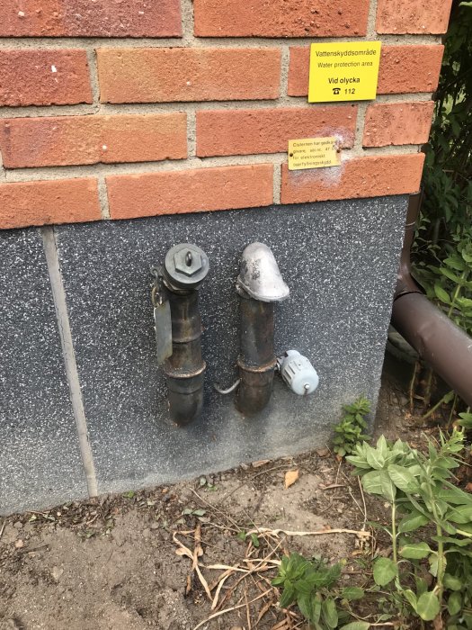 Vinkelslipade rör som sticker ut ur sockelputs vid en husvägg med tegelfasad och varningsskyltar.