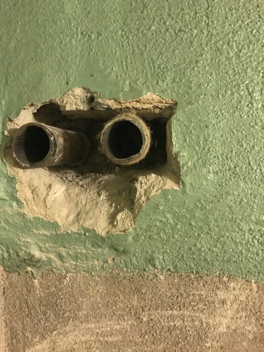 Två rör som sticker ut ur en sockelputsad vägg med synliga skador runt rören där puts har tagits bort.