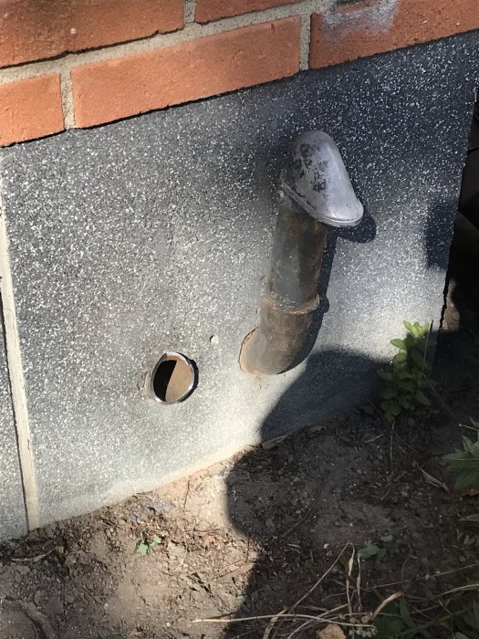Delvis kapat metallrör sticker ut från en sockelputsad vägg bredvid ett borrat hål och bakomgrunden består av tegel och markvegetation.