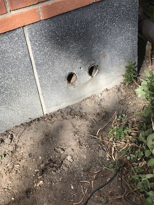Två hål i en husgrund med synlig sockelputs och rör som sticker ut, nära marknivå och vegetation.