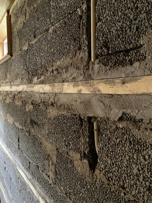 Grovt betongvägg med en horisontell träregel ingjuten och delvis utsatt, med synliga plaströr.