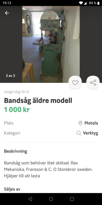 Äldre modell av bandsåg i grön färg i en verkstadslokal med bakgrund av verktyg på väggen.