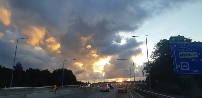 Kvällshimmel med dramatiska moln över en trafikerad motorväg med vägskyltar.