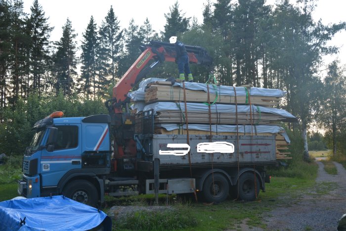 Leverans av massivt furugolv med en person på flakbilen som säkrar lasten i lantlig miljö.