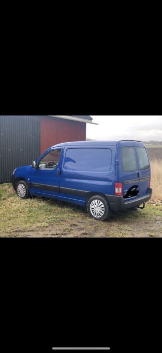 Blå renoveringsbil parkerad framför en röd lada med grå corrugated sidor på ojämn mark.