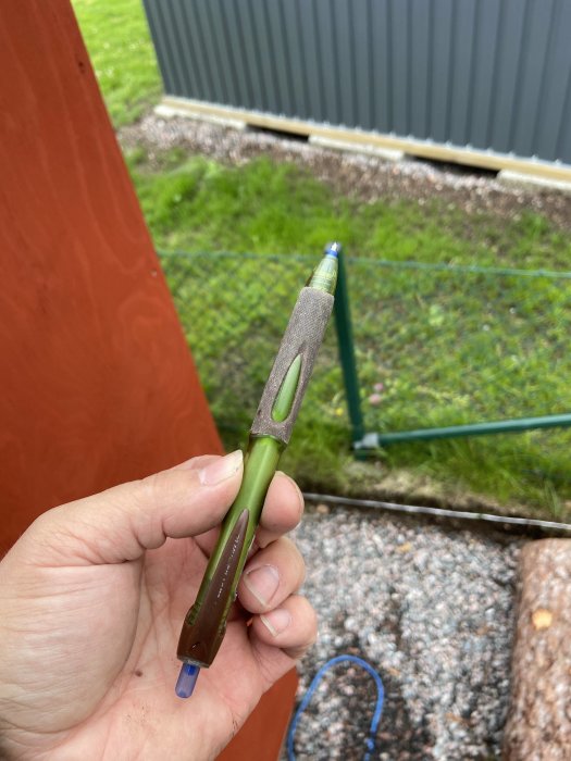 Hand håller en grön och brun Power Tank-penna med blått bläck mot en suddig bakgrund av gräsmatta och staket.