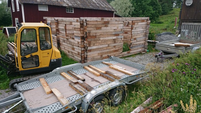 Tom biltrailer framför en minigrävare och staplar av träpallar.