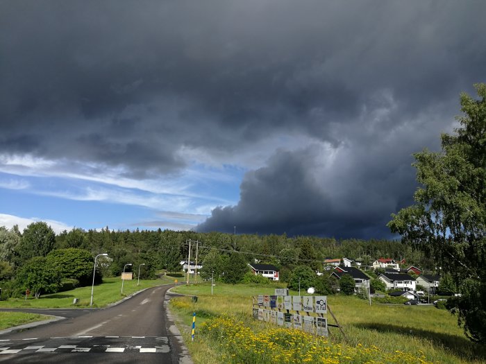 Mörka moln över ett landskap med en väg, grönska och en by med hus under en dramatisk himmel.