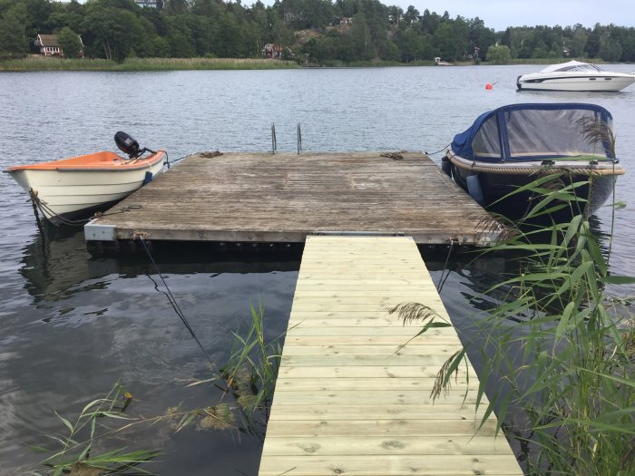 Nybyggd landgång av trä som leder till flotte vid en sjökant, omgiven av båtar och grönska.