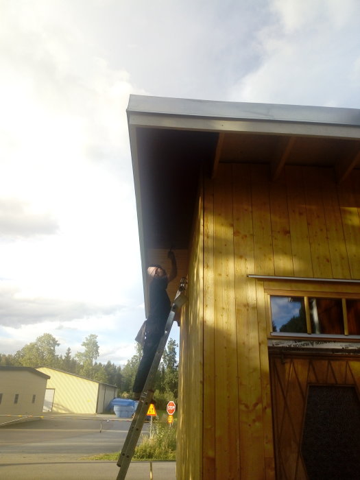 Person på stege målar takfot på trähus med rödfärg under molnig himmel.