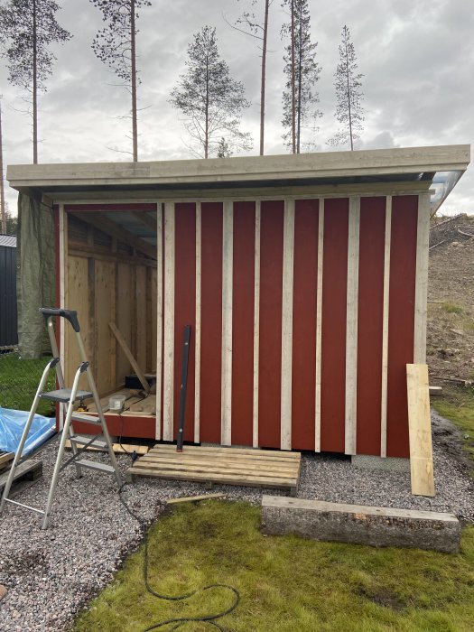 Pågående bygge av ett förråd med röda och vita vertikala plankor och ofärdig dörrfodring, omgivet av natur och byggverktyg.