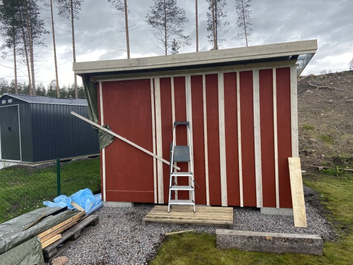 Rödmålat förråd med ofärdigt trädörrfoder och placerad stege, omgiven av skog och byggmaterial.