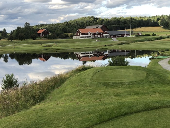 Golfbana med spegelbild av röda byggnader i vatten, grönt gräs och molnig himmel i bakgrunden.