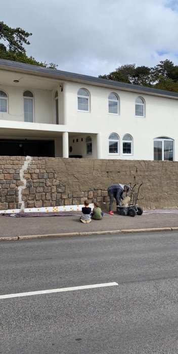 Person som arbetar med att reparera en stenmur, medan två barn sitter på gatan och tittar på.