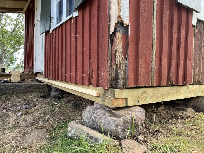 Nyinstallerad syll av trä vid husgrund med skadad rödmålad panelvägg och en katt under huset.