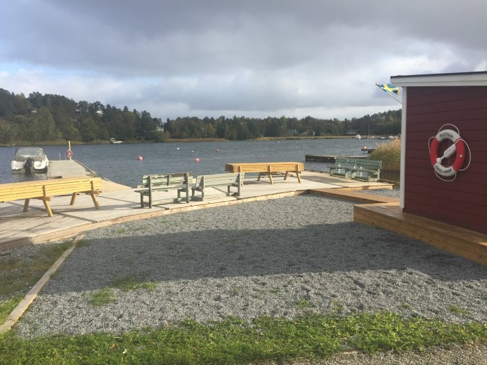 Vy över en strand med en brygga, flotte, röd livboj på en byggnad och röda nätkulor i vattnet för säkerhetsmarkering.