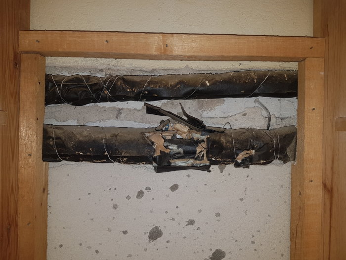Gamla rör med eventuell isolering i en väggöppning, något skadad, med omgivande träram i ett renoveringsprojekt.