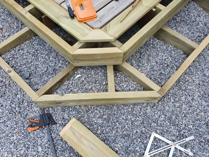 Ofärdig trätrappa med brutet hörn under konstruktion på ett grusunderlag med verktyg och brädor synliga.