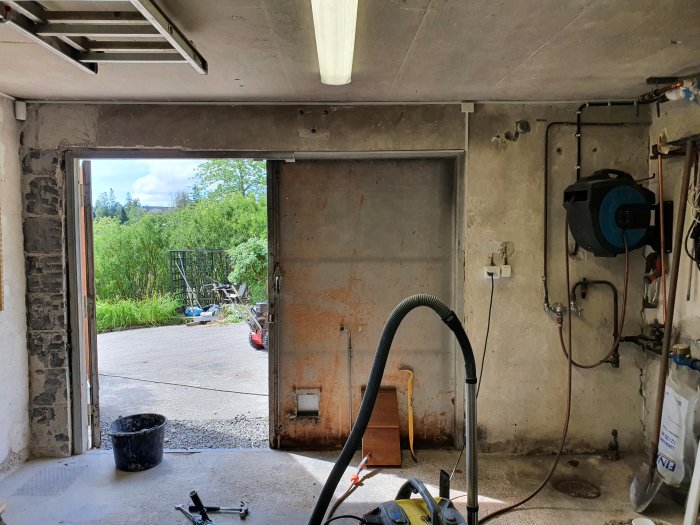 Kuva autotallista, jossa on avoin ovi ja erilaisia työkaluja ja putkia seinillä. Ulkona vehreä pihamaa ja sininen taivas.