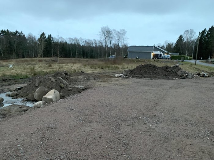 Uppgrävd mark med stora stenar och högar av jord, nära ett bostadshus i skymningen.