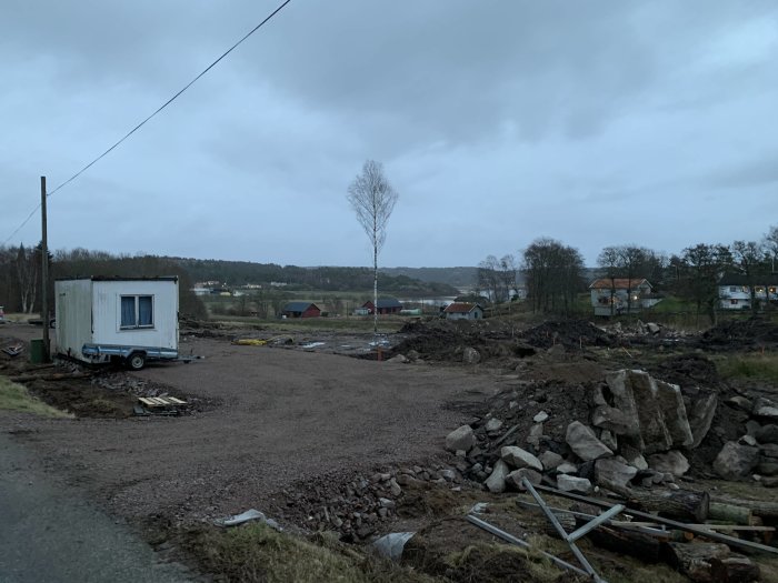 Bild av ett pågående markarbete med en byggbod, högar av grus och stenar och hus i bakgrunden under en molnig himmel.