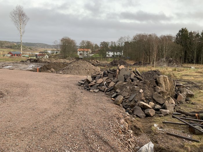 Markarbetsplats med högar av grus och sten samt uppgrävd jord under en molnig himmel.