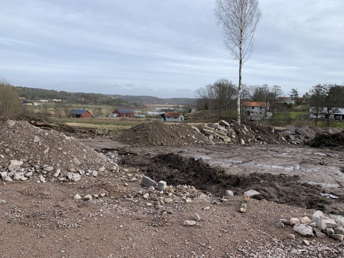 Ojämnt landskap med hopar av grus och jord vid markarbeten, omgivet av träd och byggnader i bakgrunden.