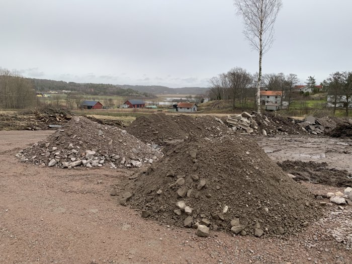 Markarbetsplats med högar av grus och rivningsavfall, trädbakgrund och byggnader i fjärran.