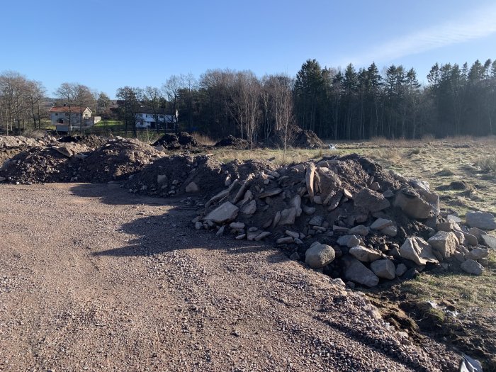 Högar av grus och stenar på en byggarbetsplats med skog och hus i bakgrunden.