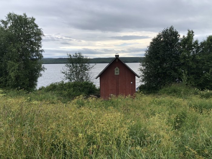 Röd stuga med vita knutar nära en sjö omgiven av grönska och träd, molnig himmel i bakgrunden.