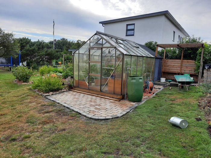 Ett växthus på en stenlagd grund med en pergola och trädgårdsverktyg i en grönskande trädgård.