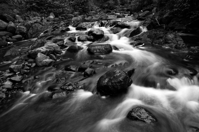 Svartvit bild av ett strömmande vattendrag med stenar och mjukt vatten i lång exponeringstid.