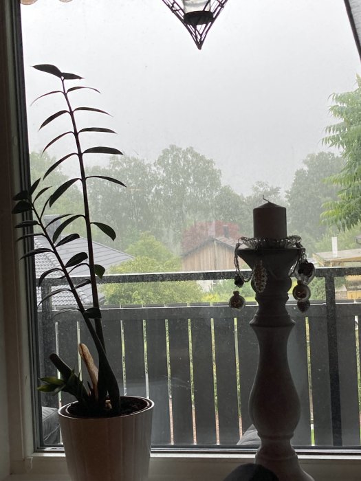 Utsikt från fönster med växt och ljusstake, synligt kraftigt regnfall och grå himmel utanför.