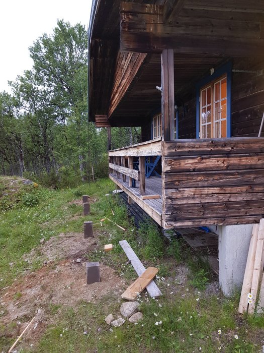 Nygrävda plintar vid en befintlig träaltan för en utbyggnad, intill ett timmerhus i en naturlig skogsmiljö.