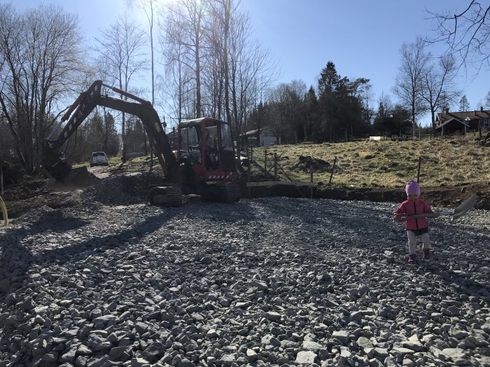 Grävmaskin och barn på en grovplanerad byggplats med grus och stubbar.