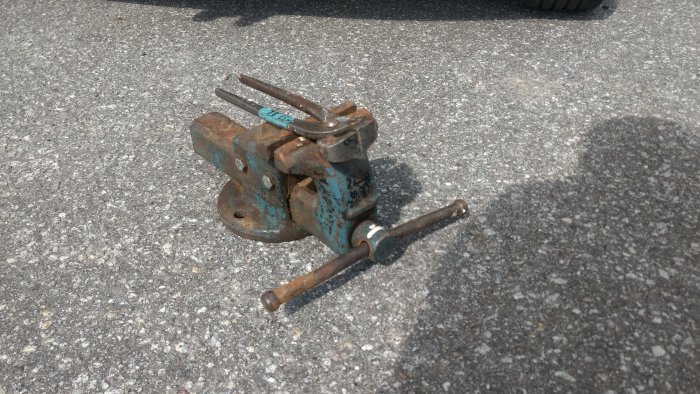 Ett gammalt rostigt skruvstäd med öppna käftar på asfalterat underlag.