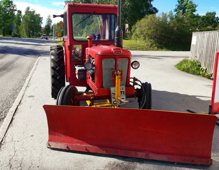 Röd veterantraktor med plog monterad fram, parkerad vid en gata på en solig dag.