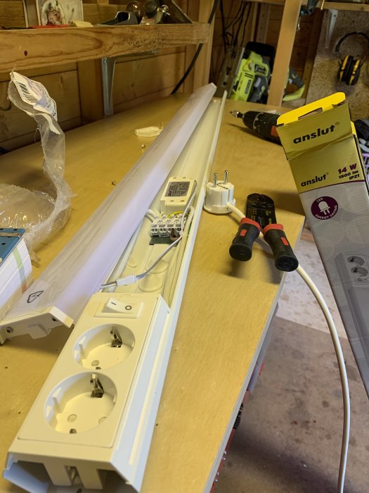 LED-lampa på arbetsbänk med dubbeluttag, förpackning och verktyg i ett verkstadsutrymme.