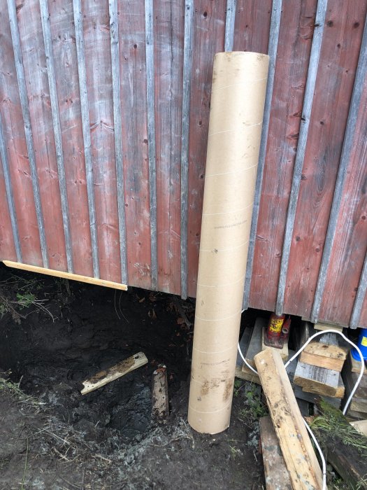 Grävd hål för fundament vid husvägg med synliga berggrund och brun kartongrör stående upp.