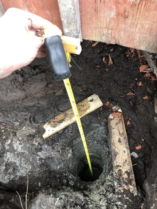 Hand håller måttband vid djupt hål i marken, visar jord och klippgrund i byggprojekt.