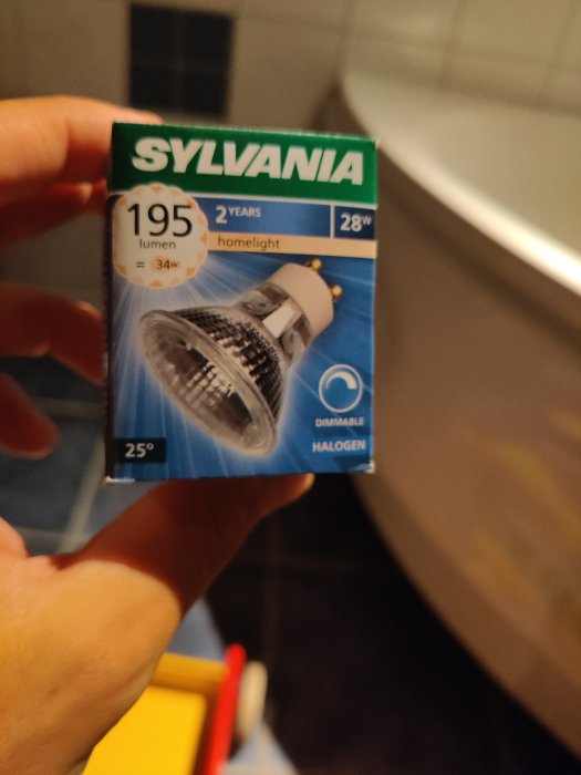 Hand håller en Sylvania halogenlampa förpackning på 28W med information om ljusstyrka och dimbarhet.