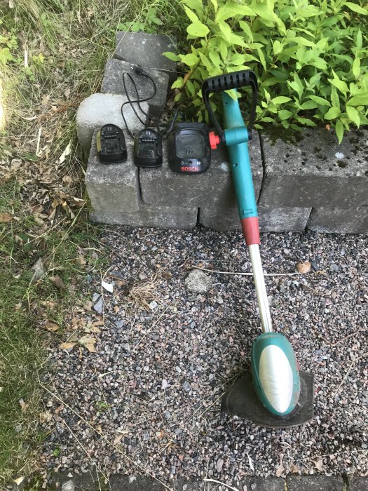 Bosch ART 26 Li batteridriven trimmer och två laddare på marken bredvid en buske.